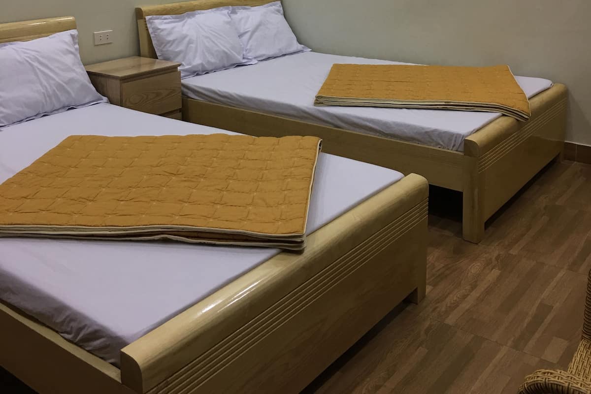 A room with two single beds, each with a wooden headboard and a white mattress. There are wooden nightstands with a small drawer and a lamp. The room has a wooden floor and a plain white wall with a light fixture above the beds.