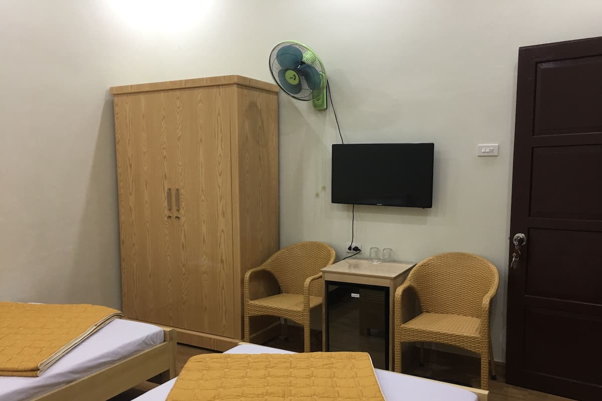 A room with two beds, a wardrobe, a TV, a fan, and two wicker chairs.