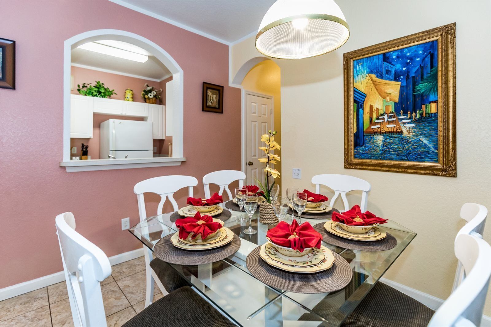 A dining area with a glass table set for six, white chairs, and a painting of a night scene on the wall.