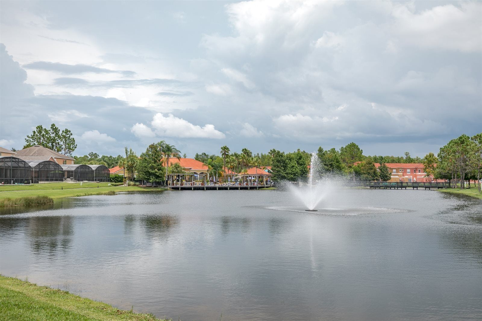 A tranquil pond with a fountain, surrounded by buildings with red roofs and green trees.