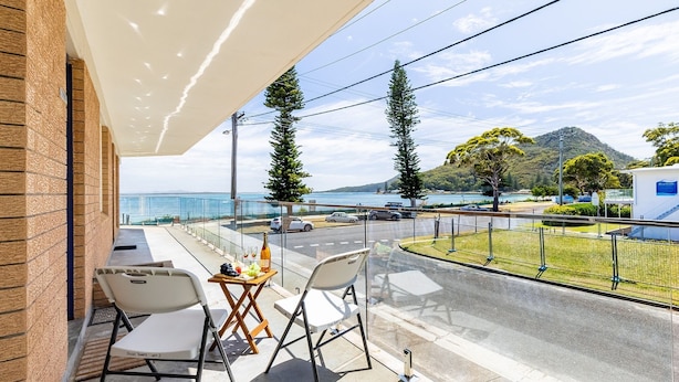 A balcony with a table set for two, overlooking a coastal road with cars and a mountain in the background. - Shoal Towers, Unit 2 11 Shoal Bay Rd Air Conditioning Wifi free Netflix water views (Shoal Bay)