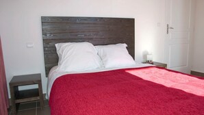 Quarto com cabeceira de madeira, roupa de cama branca, manta vermelha, mesa de cabeceira com abajur e uma porta.