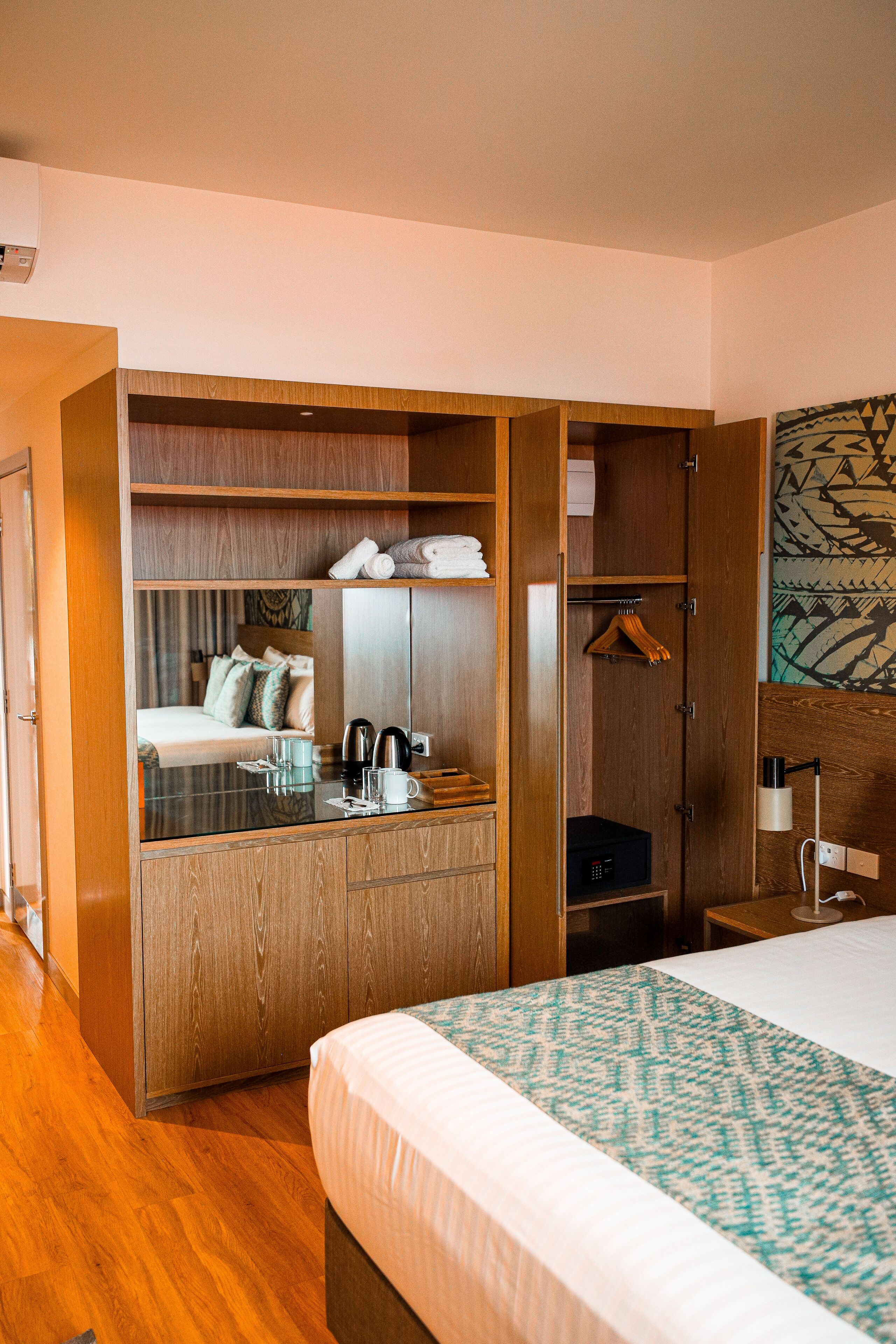A hotel room with a bed, a wardrobe, a mirror, a desk with a lamp, and a hanging coat rack.