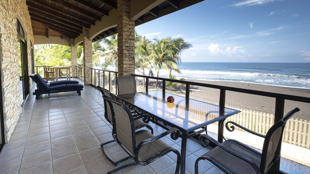 Ein überdachter Außenbereich mit Tisch und Stühlen, mit Blick auf einen Strand mit Palmen und dem Meer.