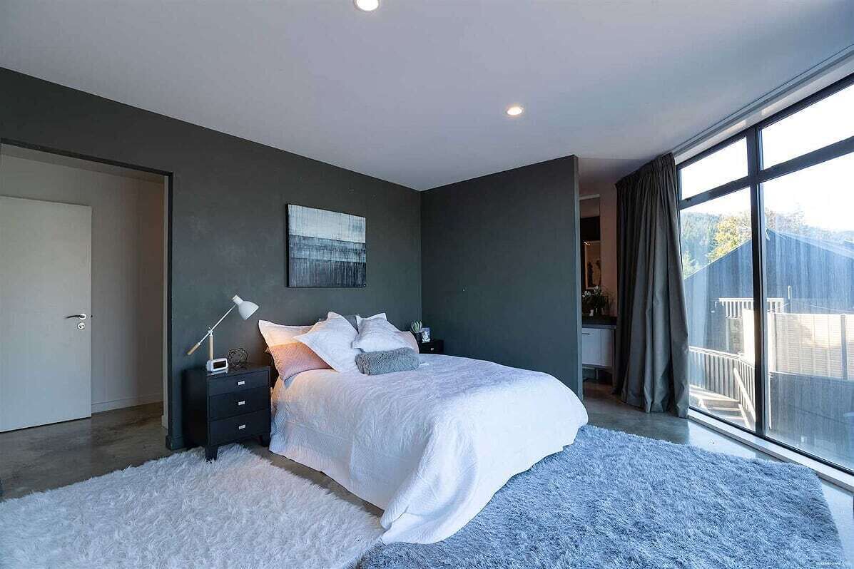 A modern bedroom with a large bed, a nightstand, a lamp, and a view of the outdoors through a large window.