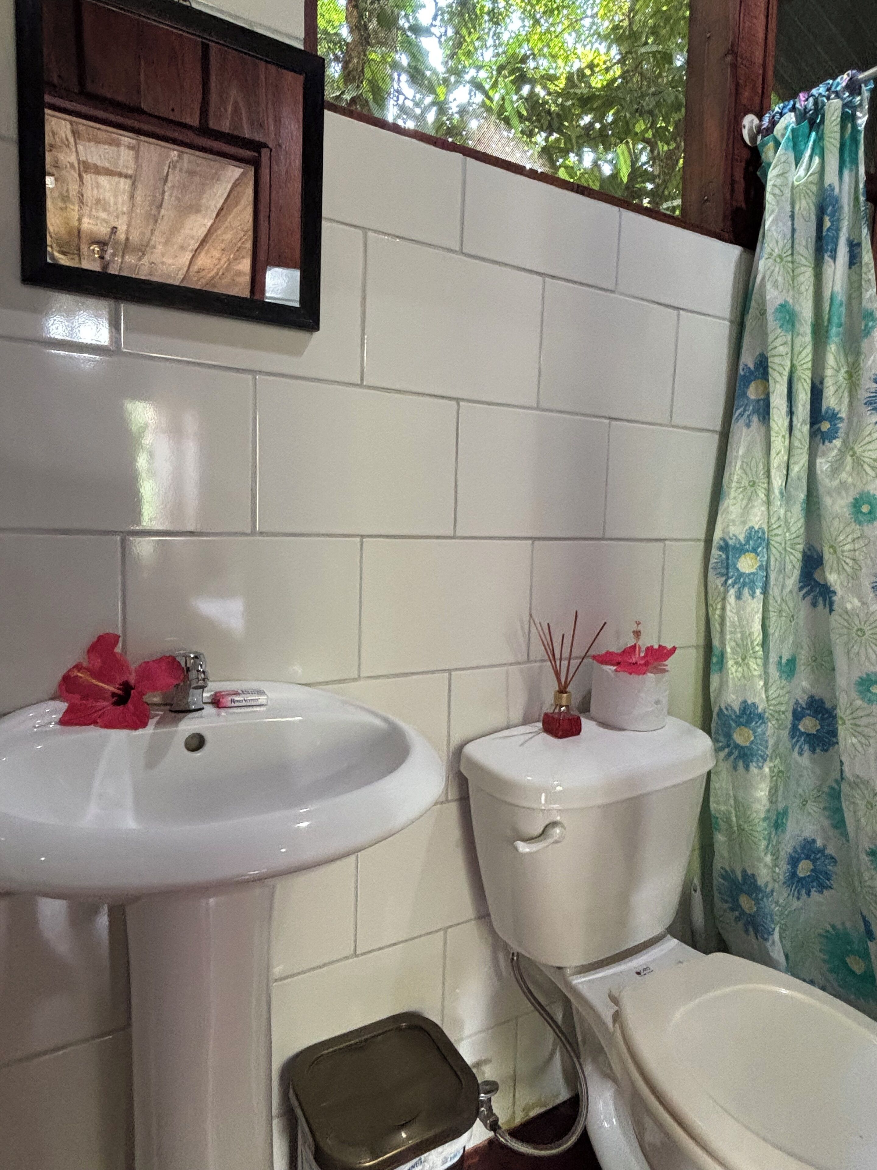 Baño con lavamanos blanco, inodoro y bidé. Hay un cubo de basura pequeño y un difusor de caña decorativo sobre el inodoro.