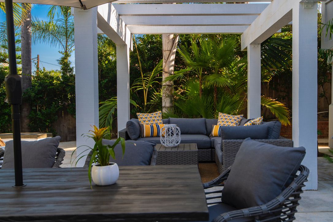 Un área de estar al aire libre con una pérgola blanca, mobiliario de mimbre y una planta en maceta.