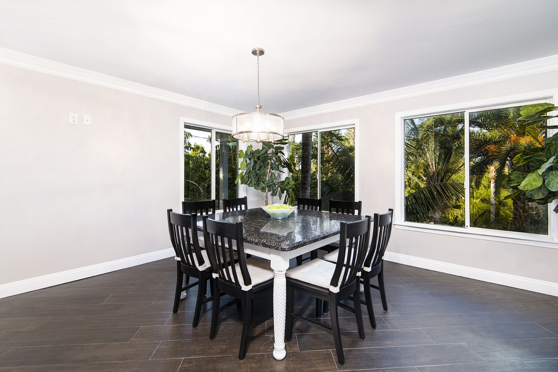 A dining area with a marble table, black chairs, a chandelier, and large windows with a view of tropical foliage.