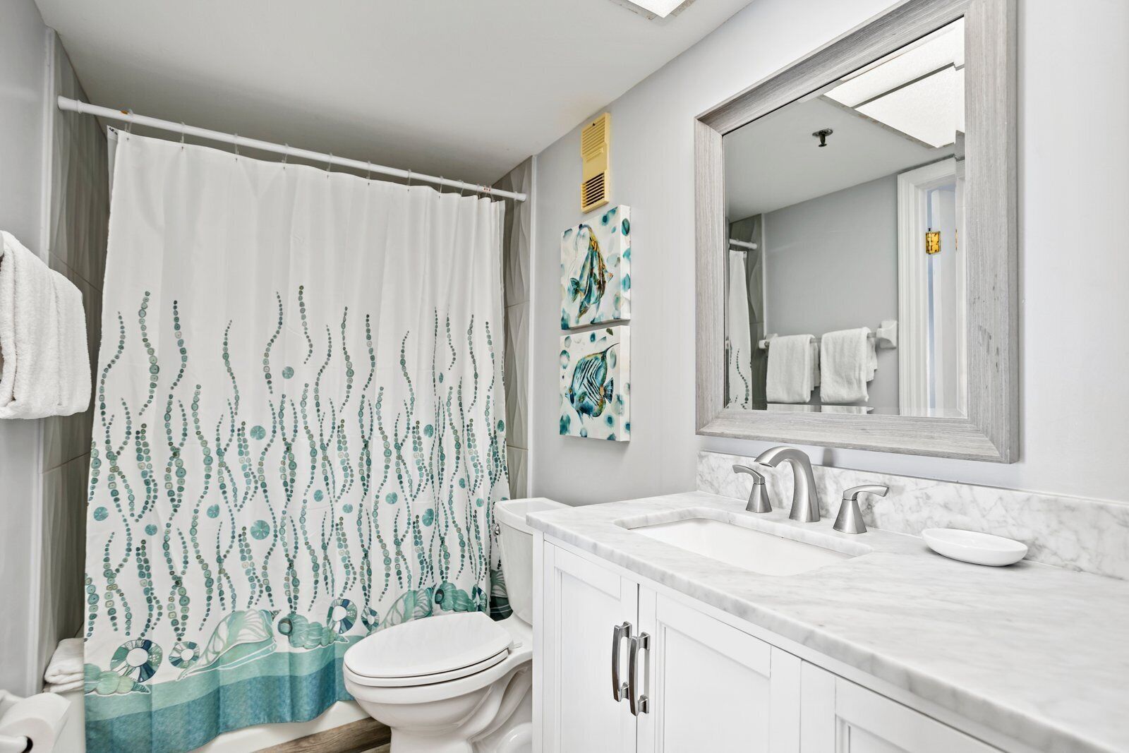 Un baño con inodoro blanco, cortina de ducha con diseño azul y verde, lavamanos blanco con encimera de mármol y espejo sobre el lavamanos.