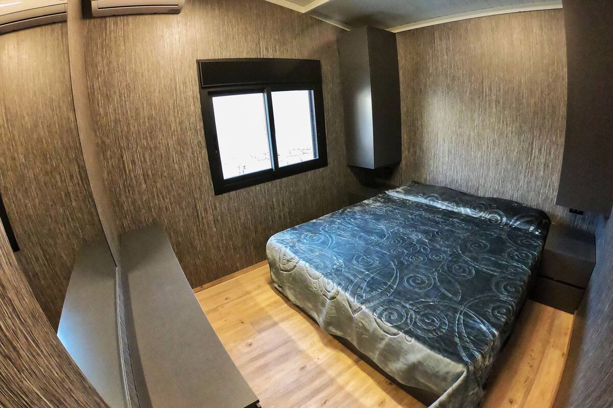 Dormitorio compacto de una caravana, con cama, una ventana pequeña y un equipo de aire acondicionado.