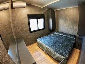 A compact RV bedroom with a bed, a small window, and an air conditioning unit. - The Getaway (Hrajel)