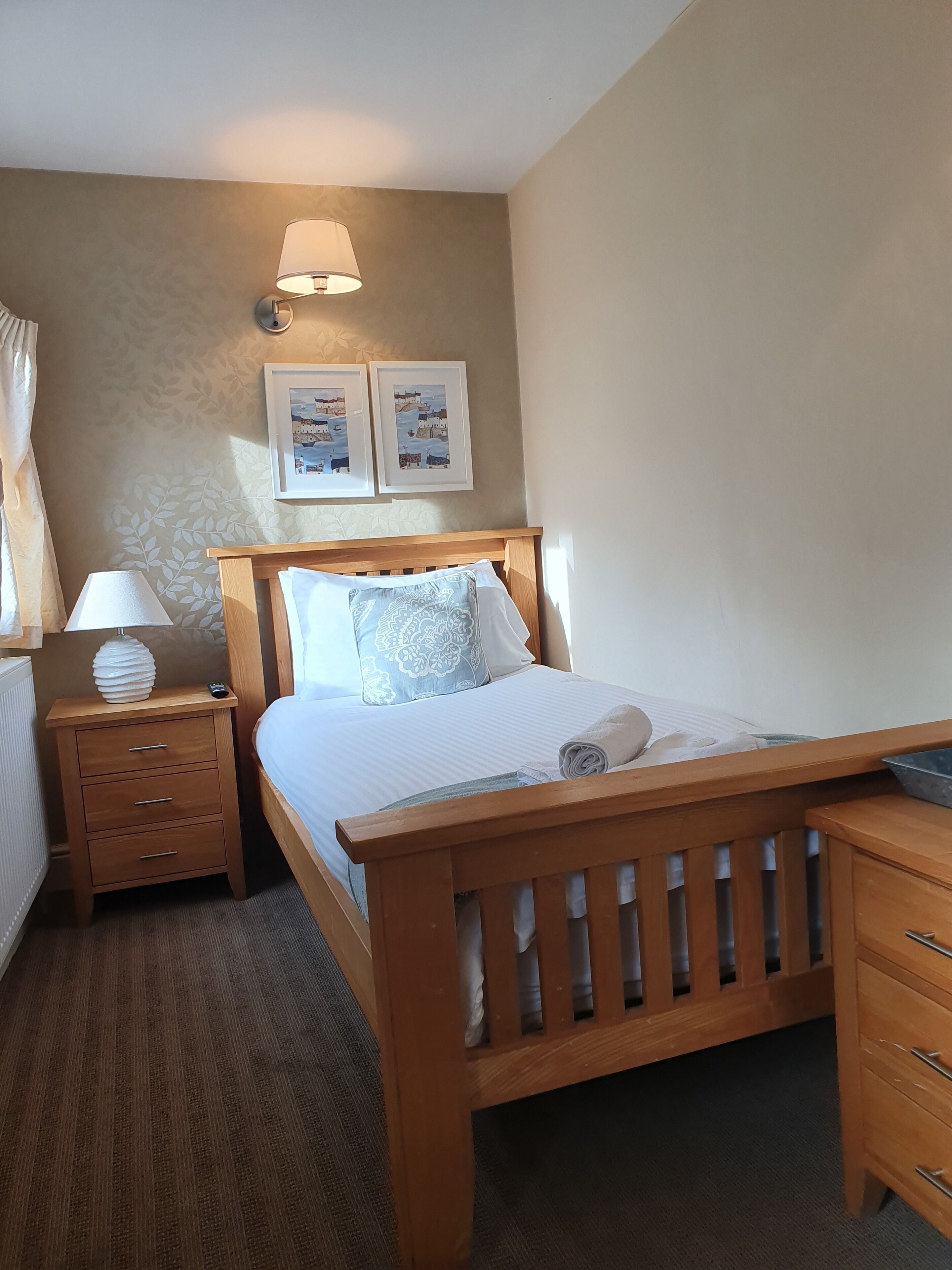Une chambre à coucher bien rangée, avec un lit en bois, une table de chevet avec une lampe, une commode et deux tableaux encadrés accrochés au mur.