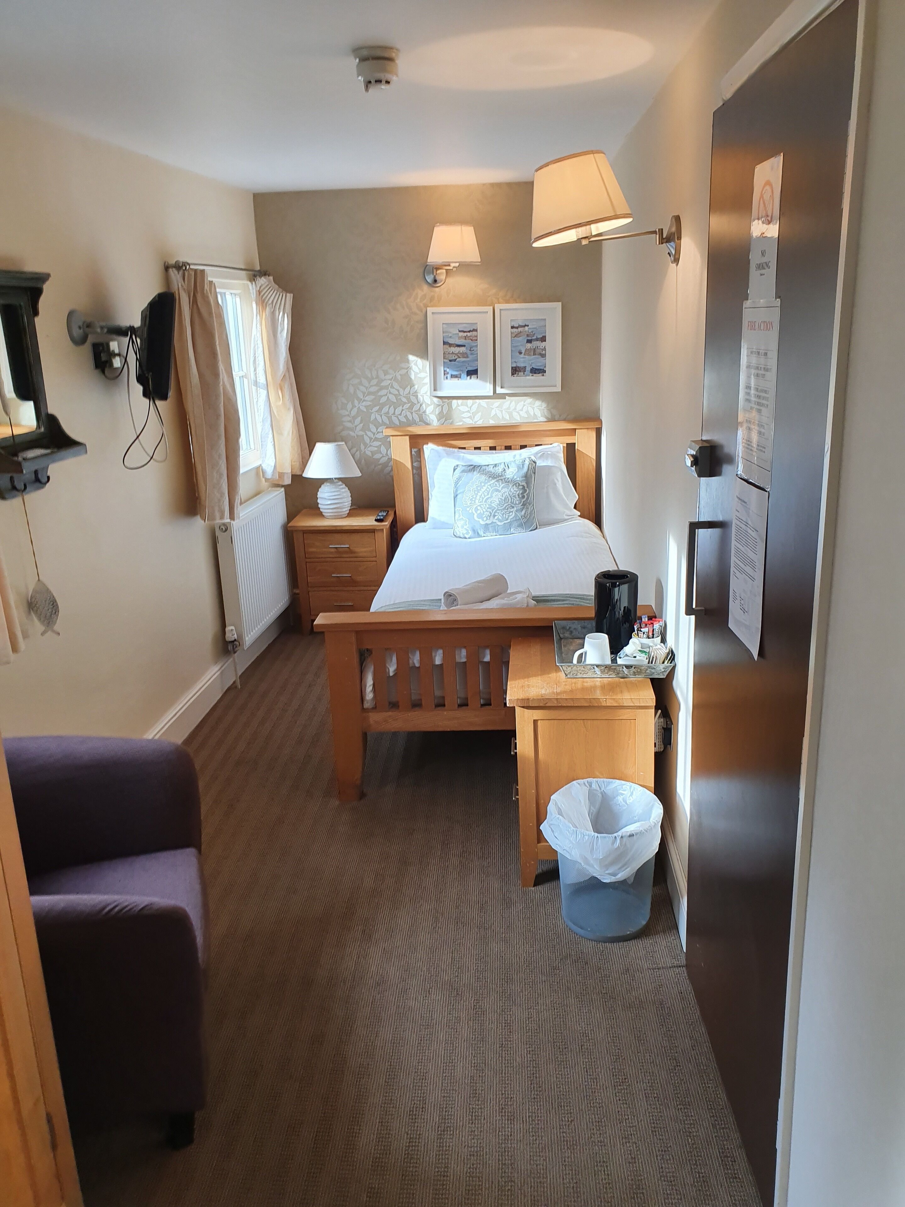 A hotel room with a bed, bedside lamps, a wooden nightstand, a small table, a trash bin, a purple chair, a radiator, a window with curtains, and a wall-mounted TV.