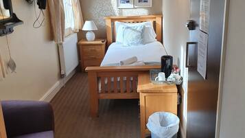 A hotel room with a bed, bedside lamps, a wooden nightstand, a small table, a trash bin, a purple chair, a radiator, a window with curtains, and a wall-mounted TV.