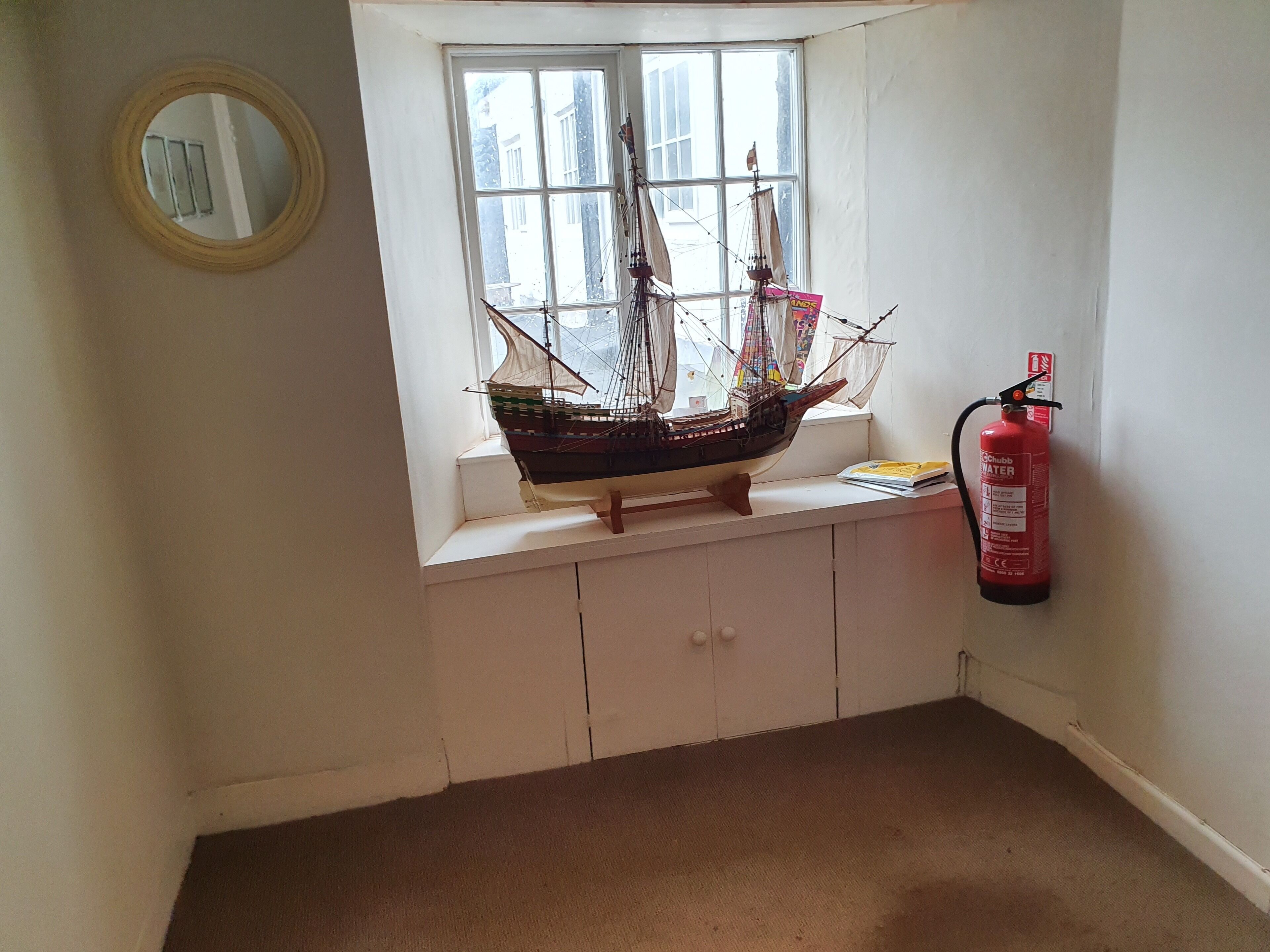 Una pequeña ventana con un modelo de barco, un extintor y un espejo en la pared.
