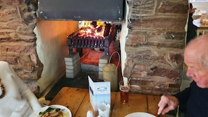 A stone fireplace with a fire burning inside, a wooden table with a plate of food, and a glass of beer. - Halfway House Inn (Torpoint)