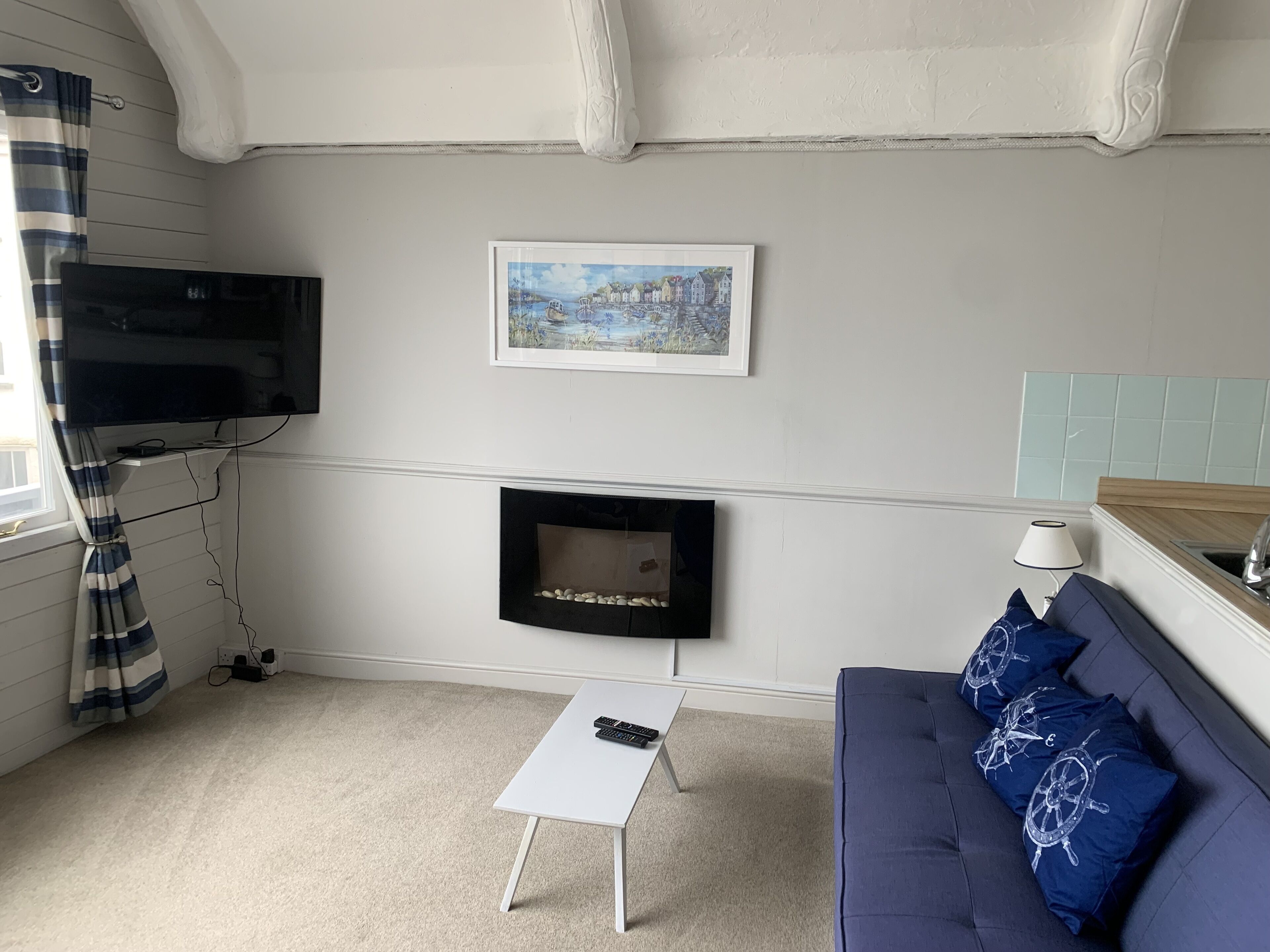 Una sala de estar con una televisión de pantalla plana montada en la pared, una chimenea debajo, una mesa de centro blanca, un sofá azul con almohadones decorativos y una ventana con cortinas.