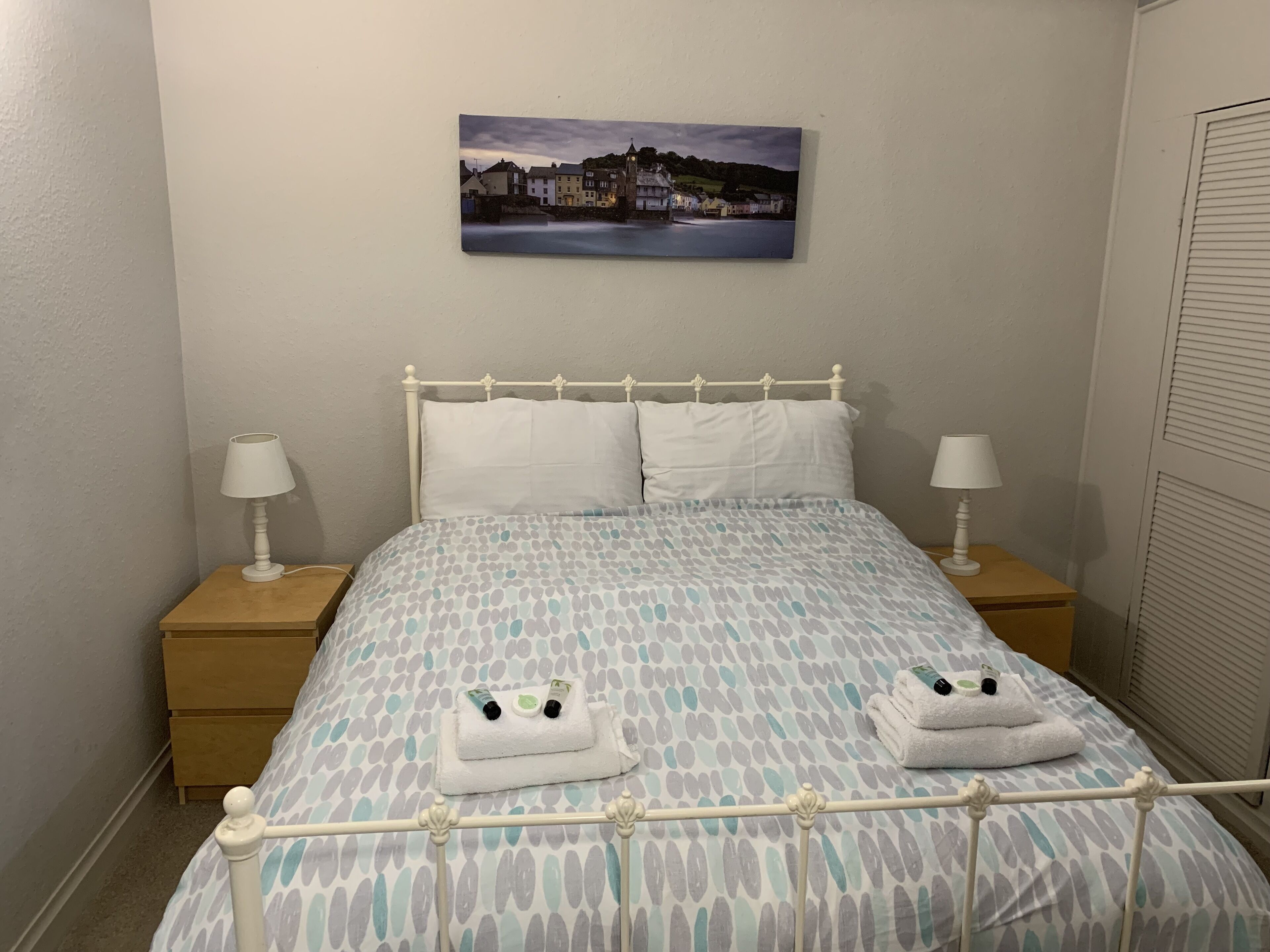 Un dormitorio con una cama, dos mesitas de noche con lámparas, un cuadro en la pared y una ventana con persianas.