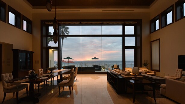 Un vaste salon offrant une vue imprenable sur l’océan au coucher du soleil, aménagé avec des canapés en cuir et un coin repas.