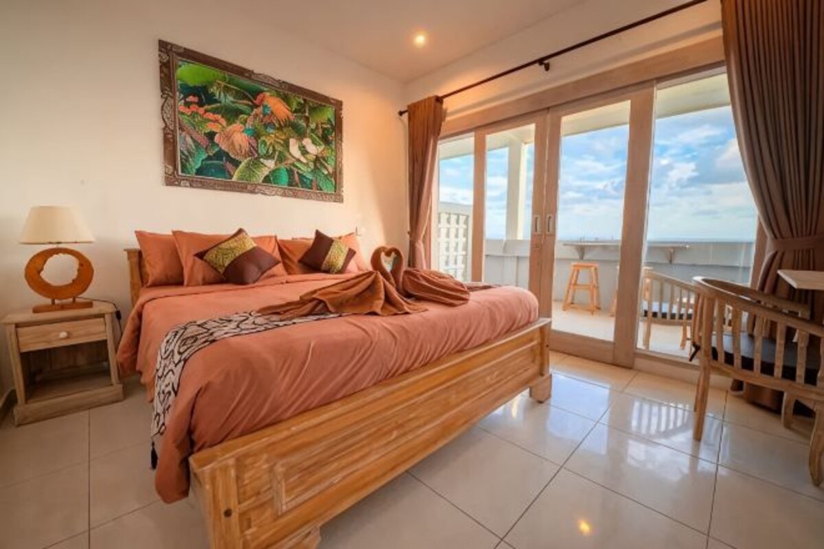 A bedroom with a bed, a nightstand, a painting, a balcony with a view, and a chair.