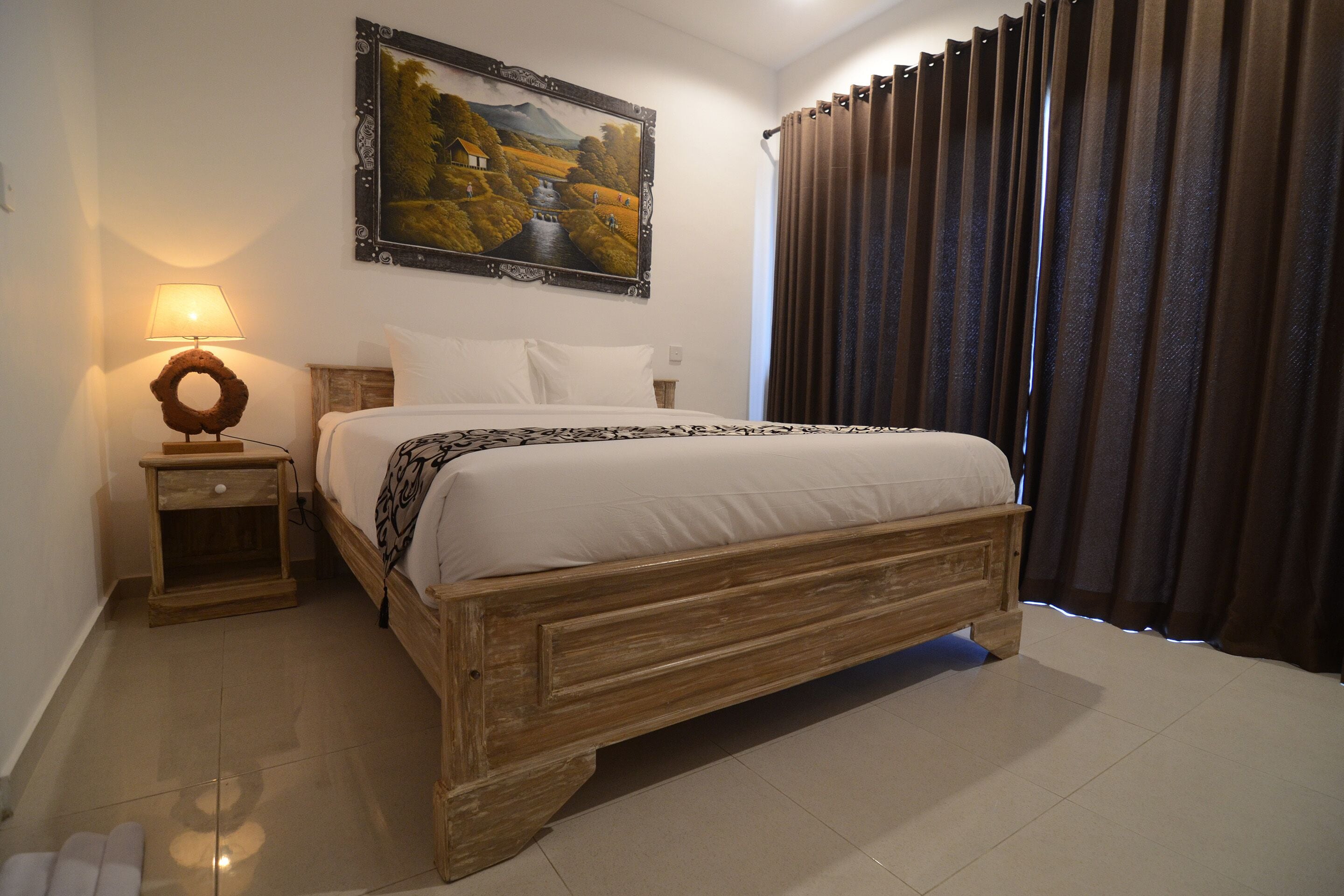 Un dormitorio con una cama de madera, una mesita de noche con una lámpara, un cuadro en la pared y una ventana con cortinas.