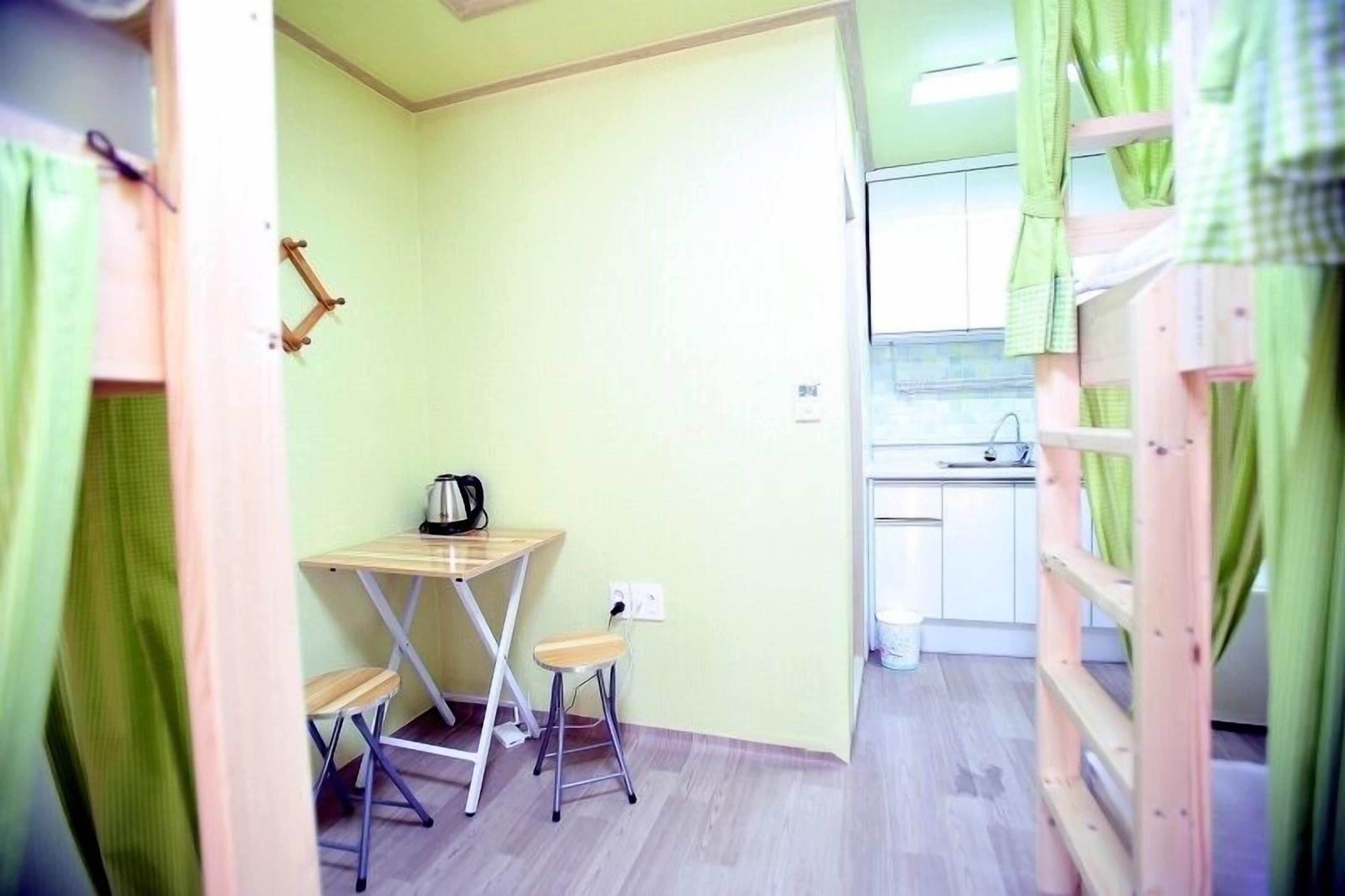 Une petite pièce comprenant une table et un tabouret, un coin cuisine avec un évier et un lit superposé avec des rideaux verts.