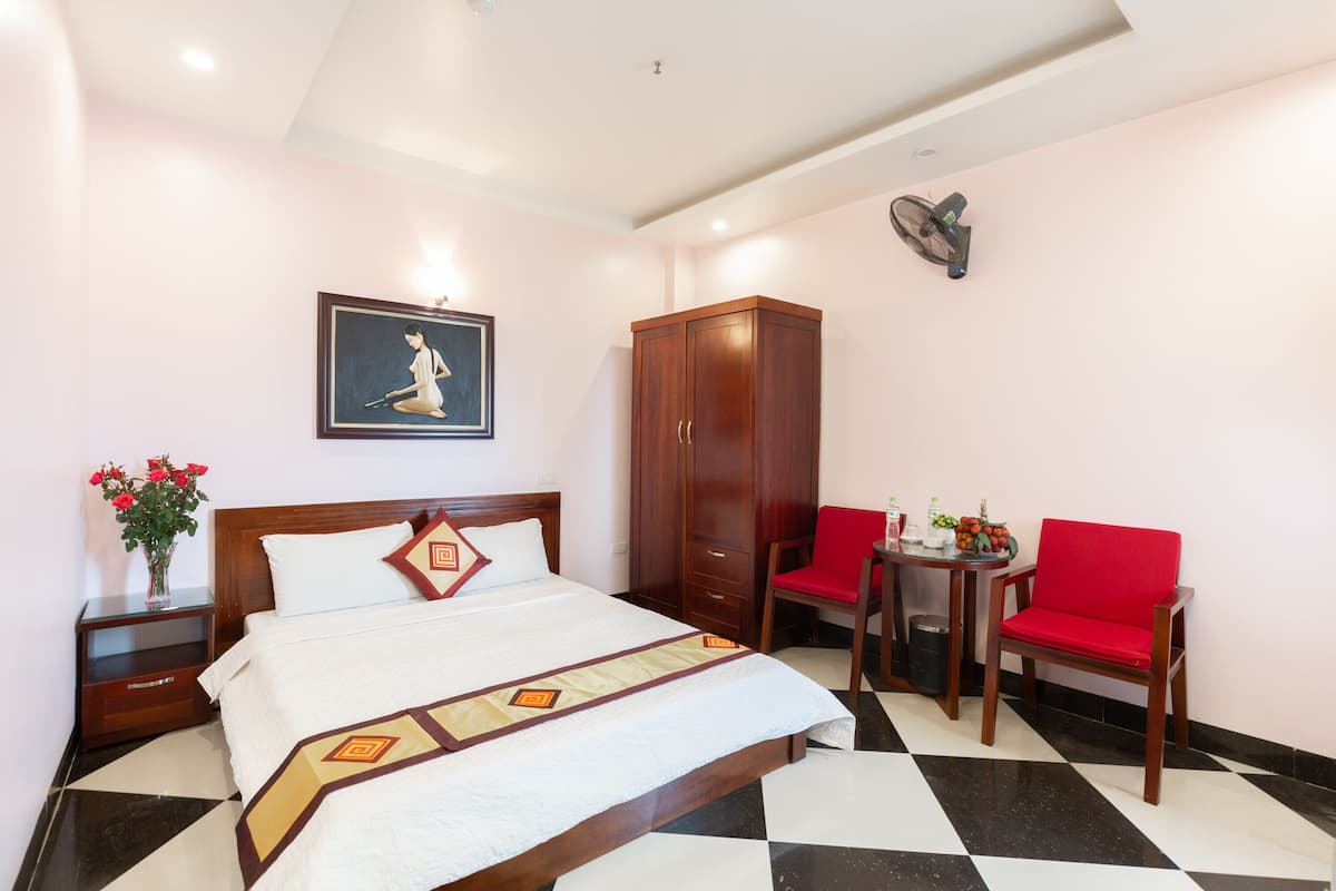 A hotel room with a bed, a wooden wardrobe, a red chair, a small table with a vase of flowers, and a ceiling fan.