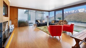 A modern living room with a wooden floor, two black leather sofas, a red armchair, and a wooden table. Large windows offer a view of mountains and a lake. - Design, Elegance and This Stunning View (Queenstown)