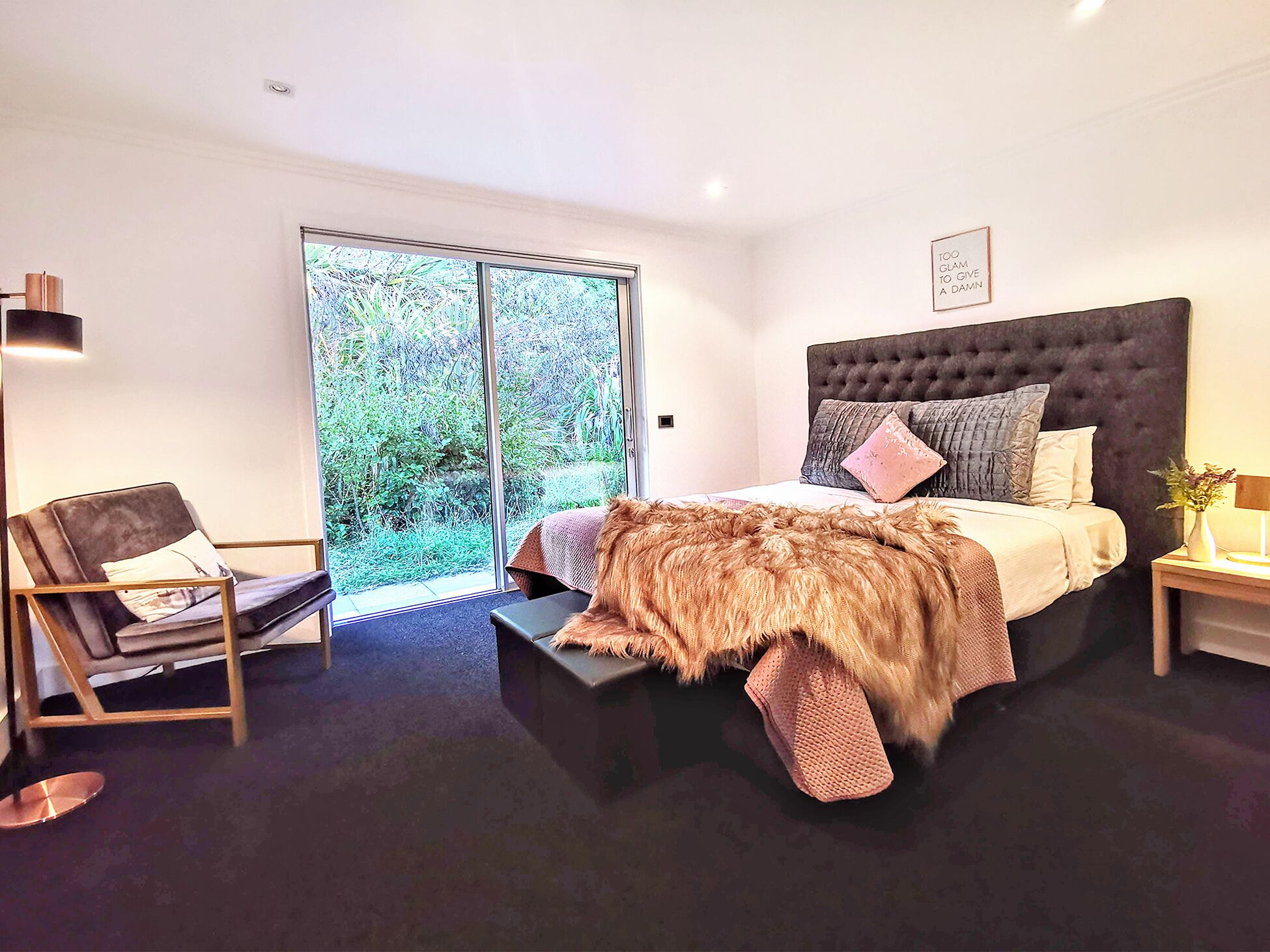 A modern bedroom with a large bed, a chair, a nightstand, and a view of greenery through a sliding door.