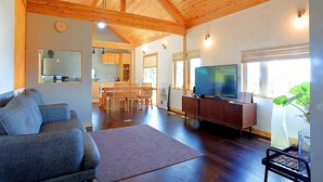 Ein geräumiges Wohnzimmer mit Holzboden, einem Fernseher, einem Essbereich und einer durchgehenden Tür erkennbaren Küche.
