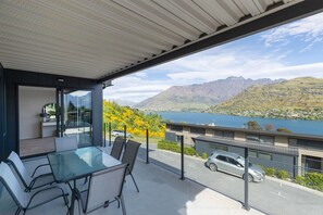 Un balcon extérieur moderne, doté d’une table et de chaises en verre, offrant une vue sur un lac et des montagnes.