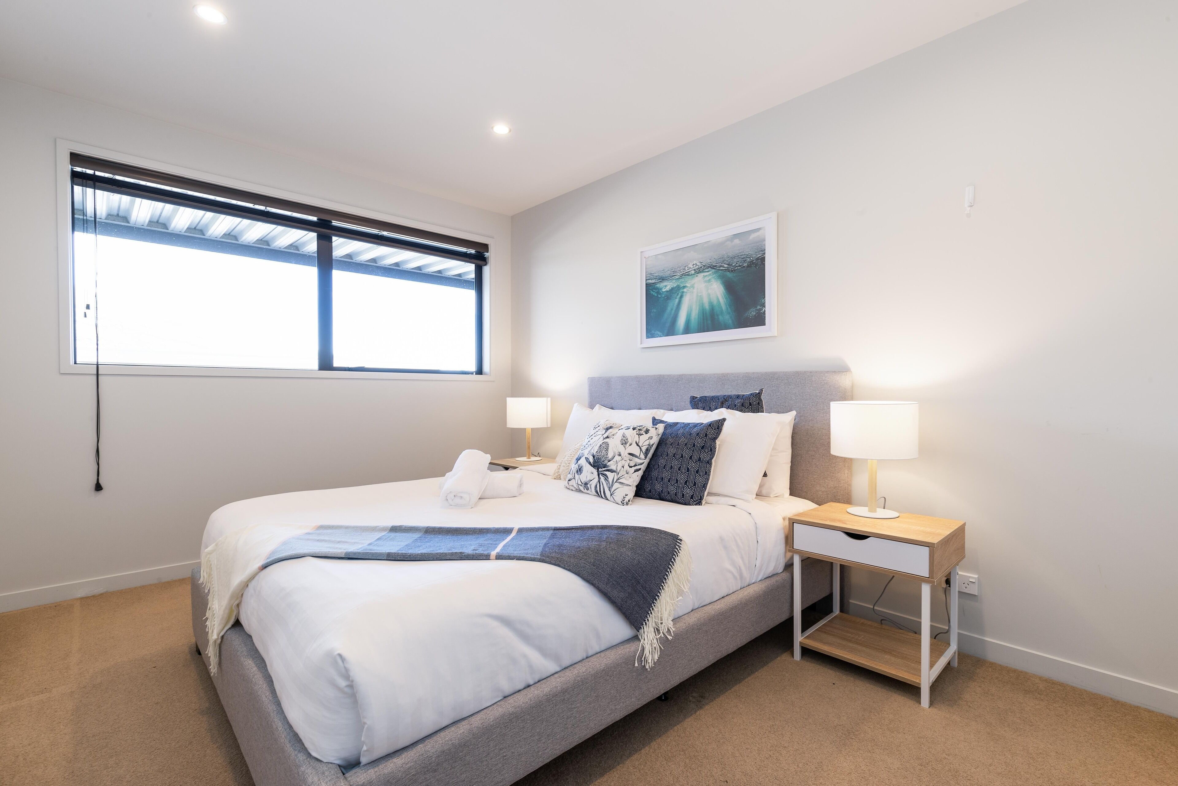 A modern bedroom with a large bed, bedside tables with lamps, a window with blinds, and a wall-mounted artwork.