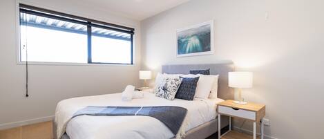 A modern bedroom with a large bed, bedside tables with lamps, a window with blinds, and a wall-mounted artwork.