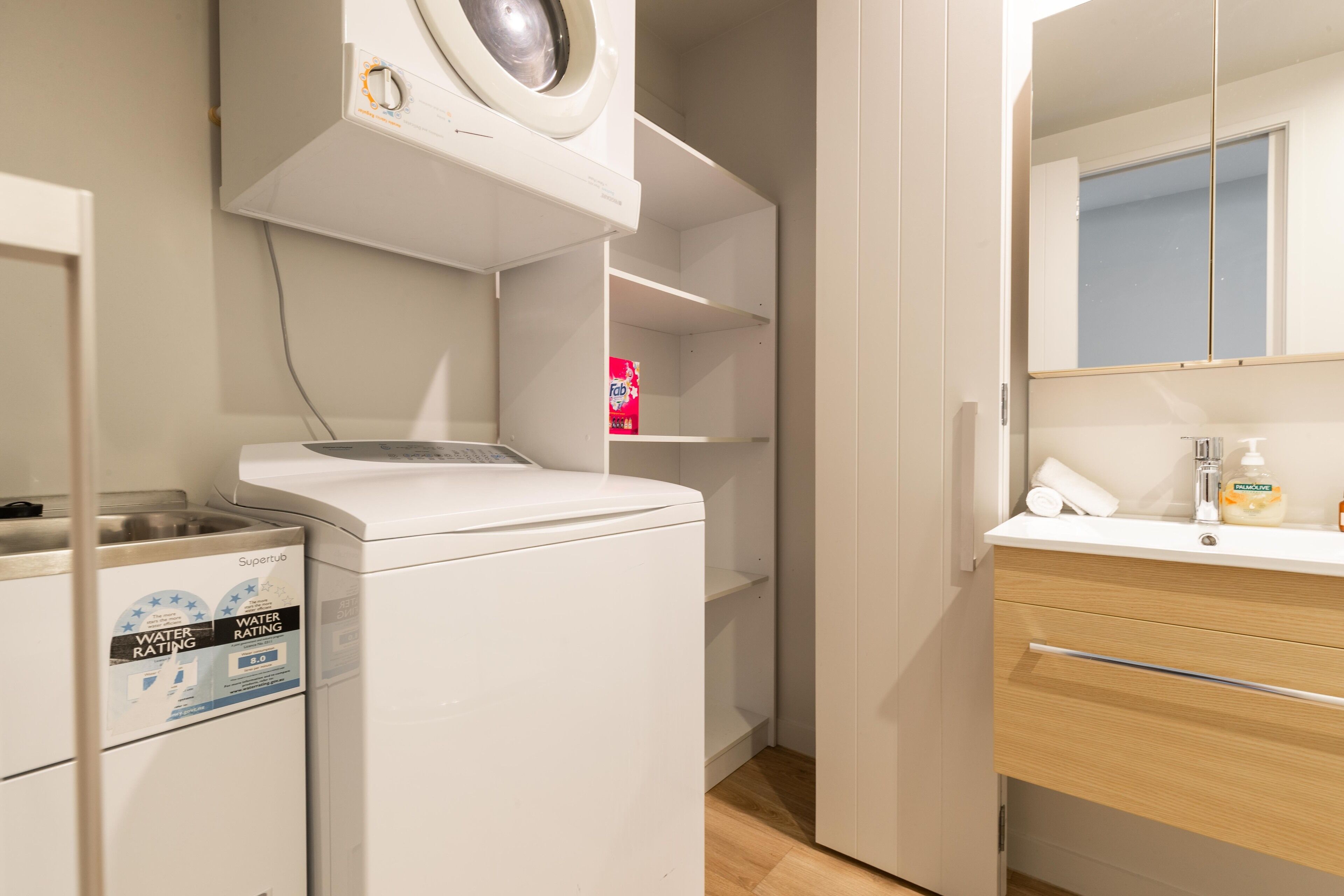 A compact laundry area with a washing machine and dryer, a small shelf with a detergent box, and a bathroom with a mirror and toiletries.