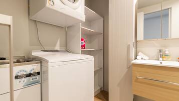 Un área de lavandería compacta con lavadora y secadora, un estante pequeño con caja de detergente, y un baño con espejo y artículos de aseo.