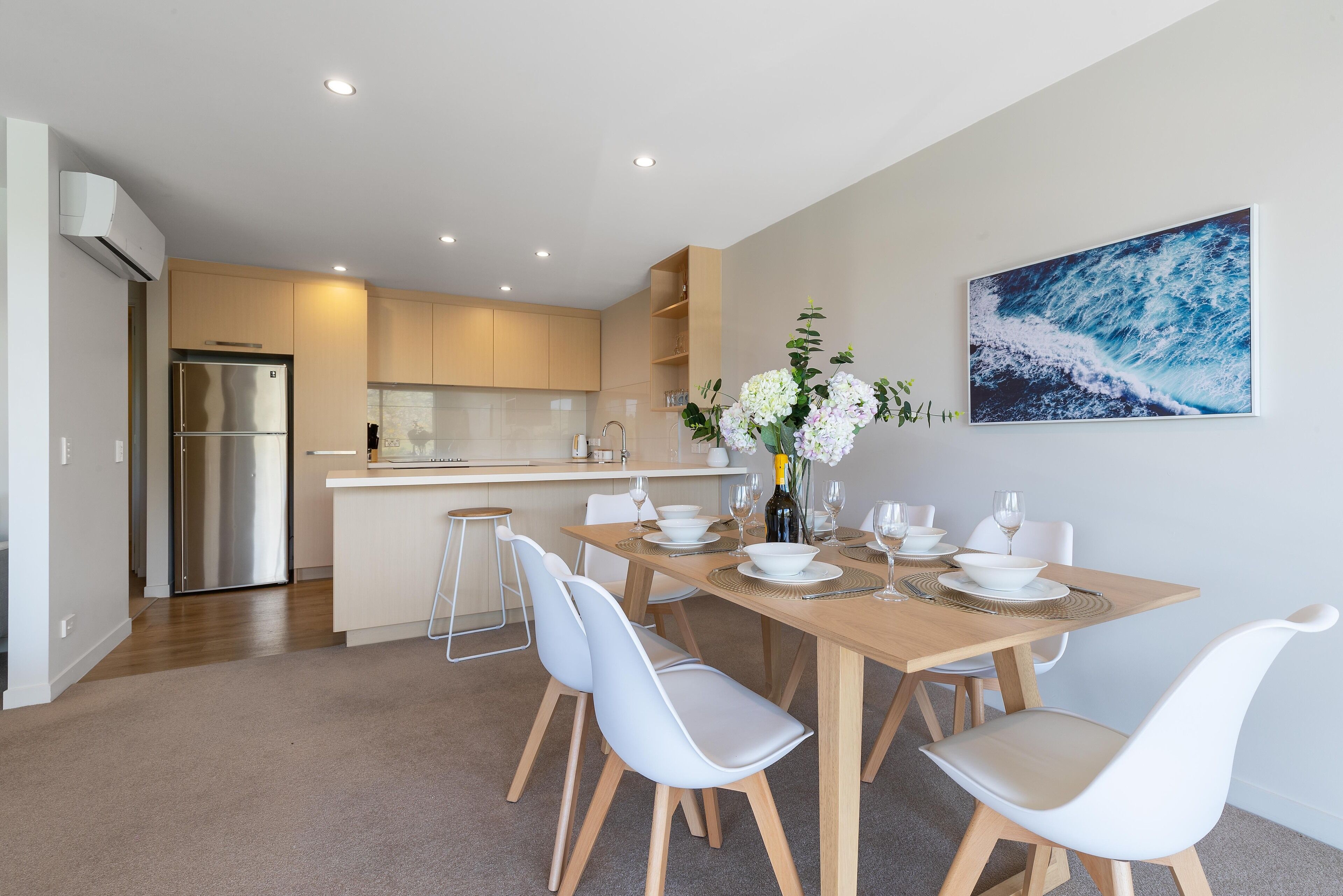 Une cuisine moderne dotée d’un réfrigérateur encastré, un coin repas avec une table en bois et des chaises blanches, et un grand tableau abstrait accroché au mur.