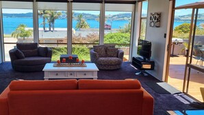 Ein Wohnzimmer mit einer Couch, einem Sessel, einem Hocker, einem Couchtisch und einem Fernseher. Große Fenster bieten Blick auf das Meer und Grünflächen.