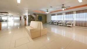 Un hall amplio con un sofá color beige, sillas blancas, un ventilador de techo y ventanas grandes con cortinas.