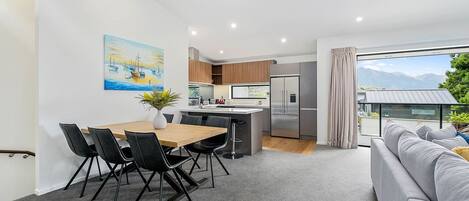 A modern kitchen with a central island, a dining area with a wooden table and chairs, and a living area with a sofa and a large window offering a view of the mountains.