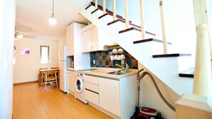 A compact kitchen with a washing machine, a dining area with a table and chairs, and a wooden staircase. - Eltz Town Pension (Jeju City)