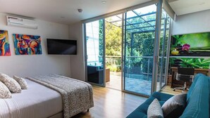 A modern bedroom with a bed, a sofa, a TV, and a sliding glass door leading to an outdoor area with a patio and greenery. -  Libertg Hotels Spa Medellin (Copacabana)