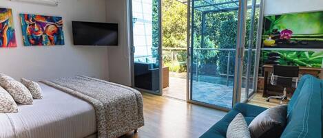 A modern bedroom with a bed, a sofa, a TV, and a sliding glass door leading to an outdoor area with a patio and greenery.