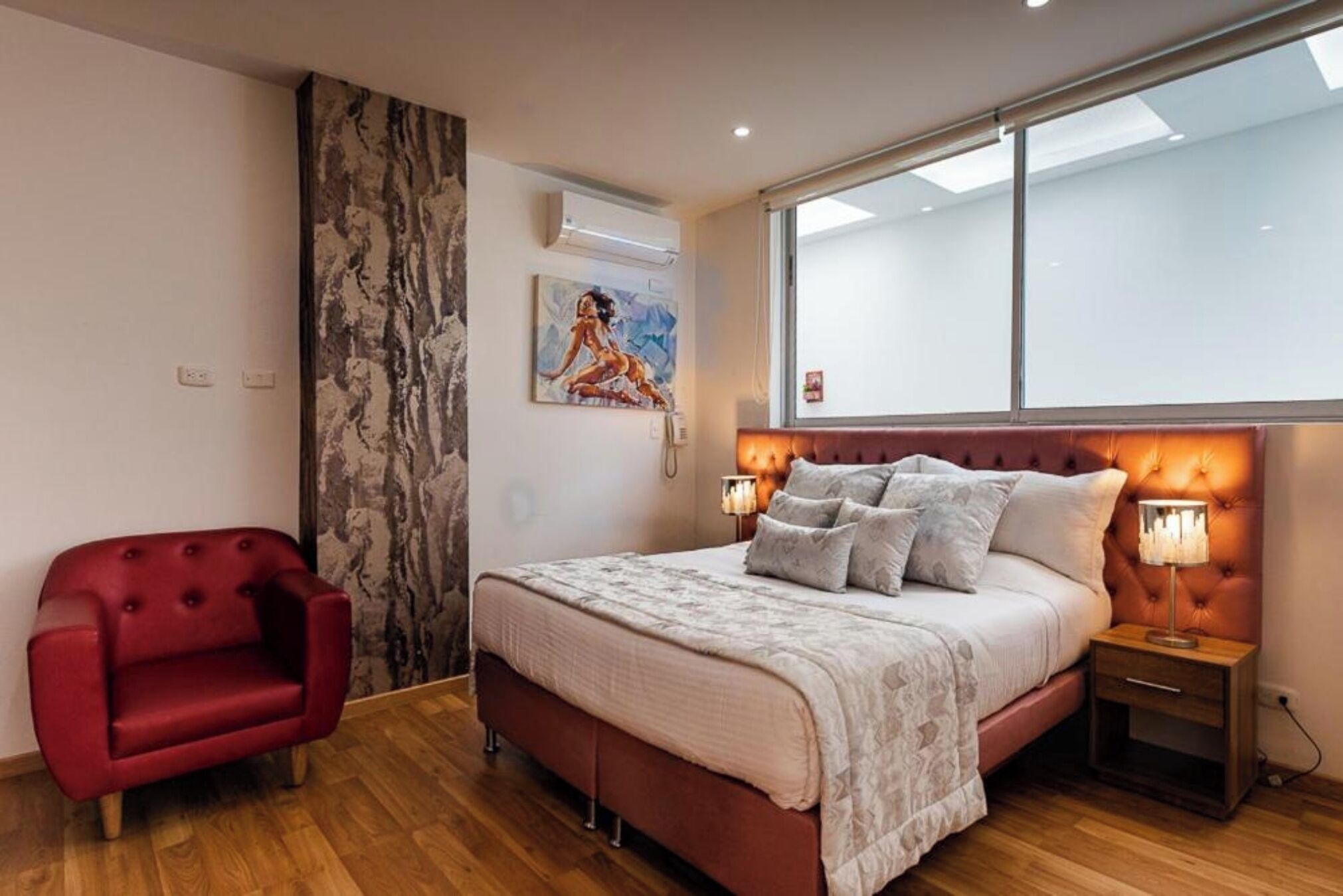 Un dormitorio con una cama grande, un sillón rojo, un cuadro en la pared y una ventana con vistas.