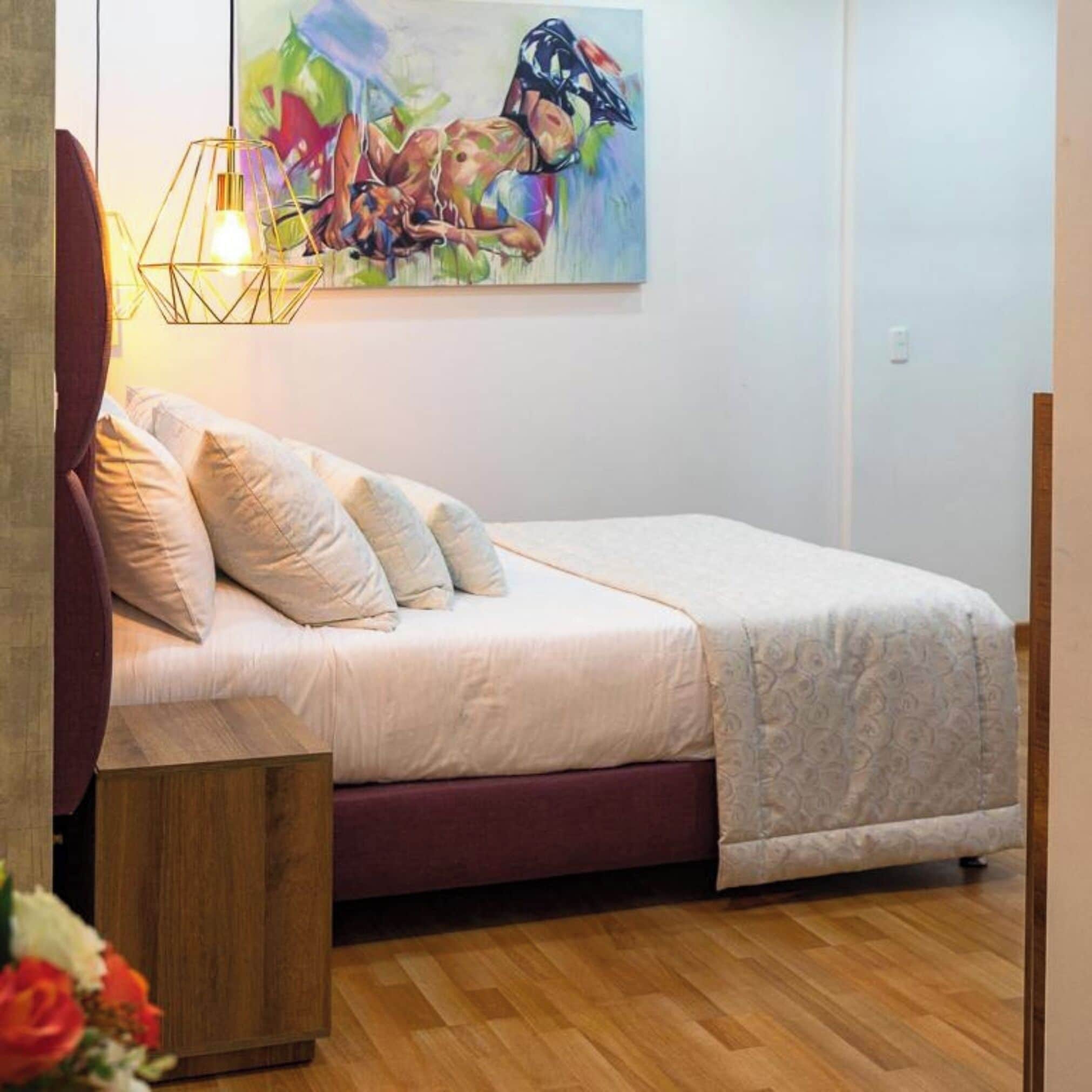 Une chambre à coucher avec un lit, des oreillers, une table de chevet en bois, un tableau accroché au mur et un luminaire suspendu.