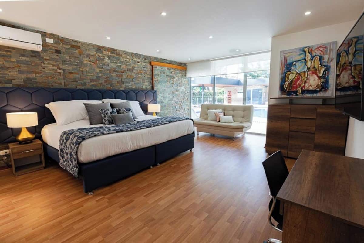 A modern bedroom with a large bed, a sofa, a wooden desk, and a stone wall.