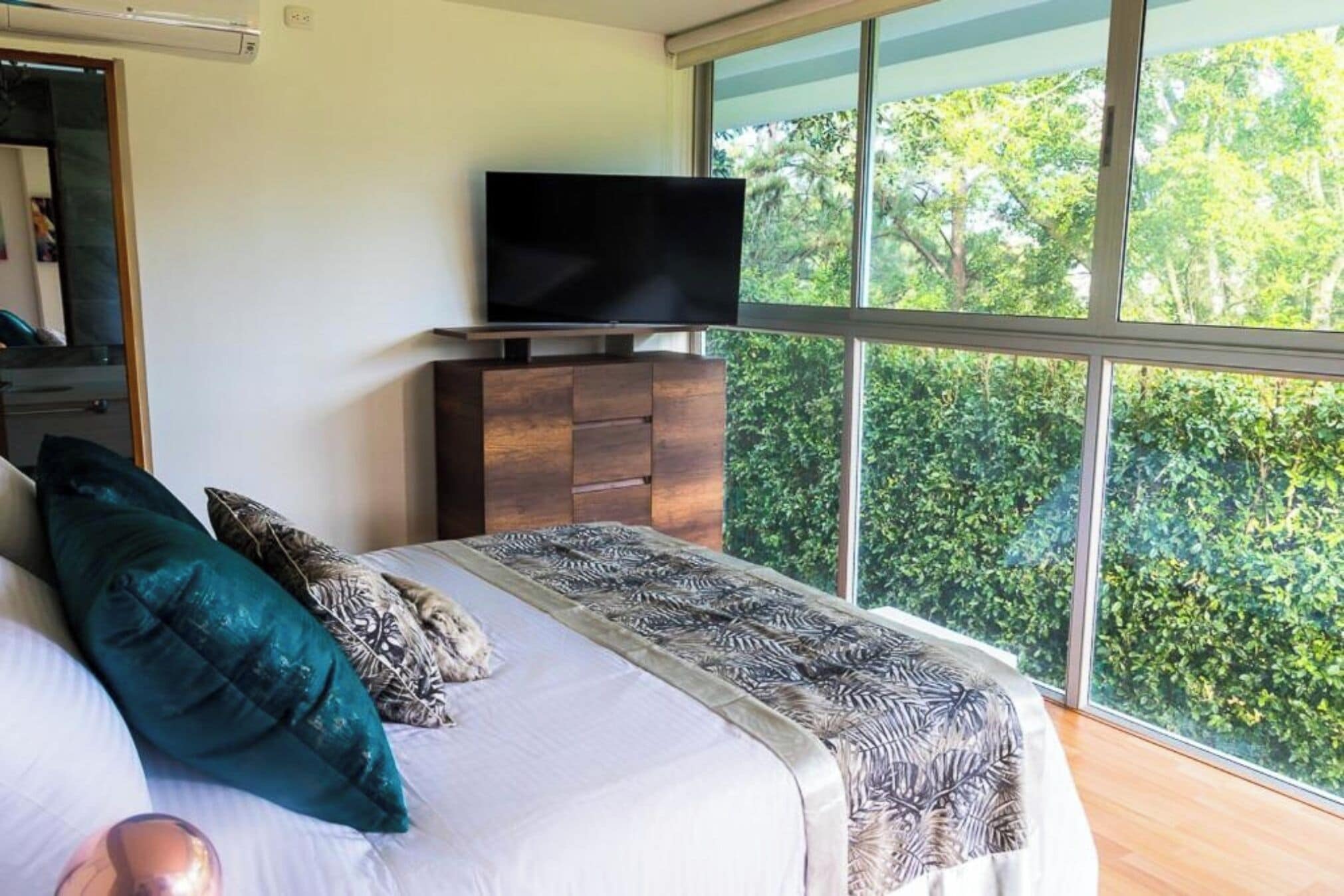 A bedroom with a large window, a flat-screen TV on a wooden cabinet, and a bed with patterned bedding.