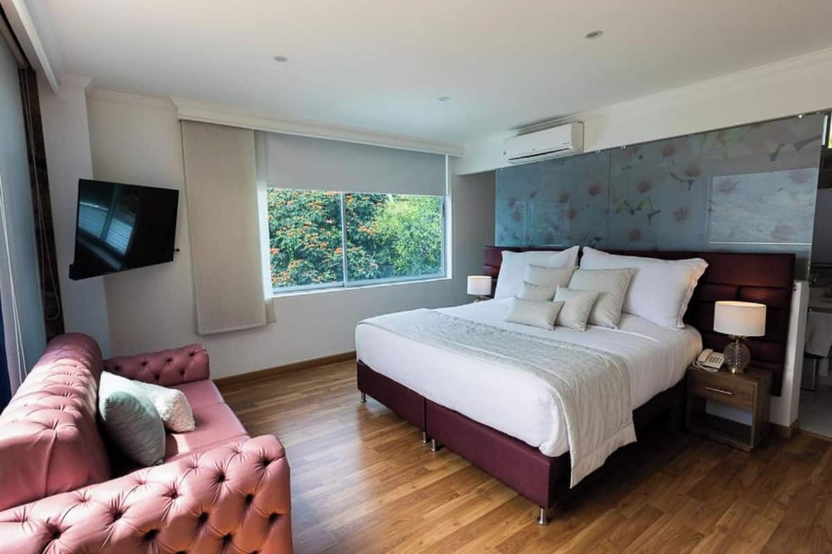 A modern bedroom with a large bed, a pink sofa, a TV mounted on the wall, and a window with a view of greenery.