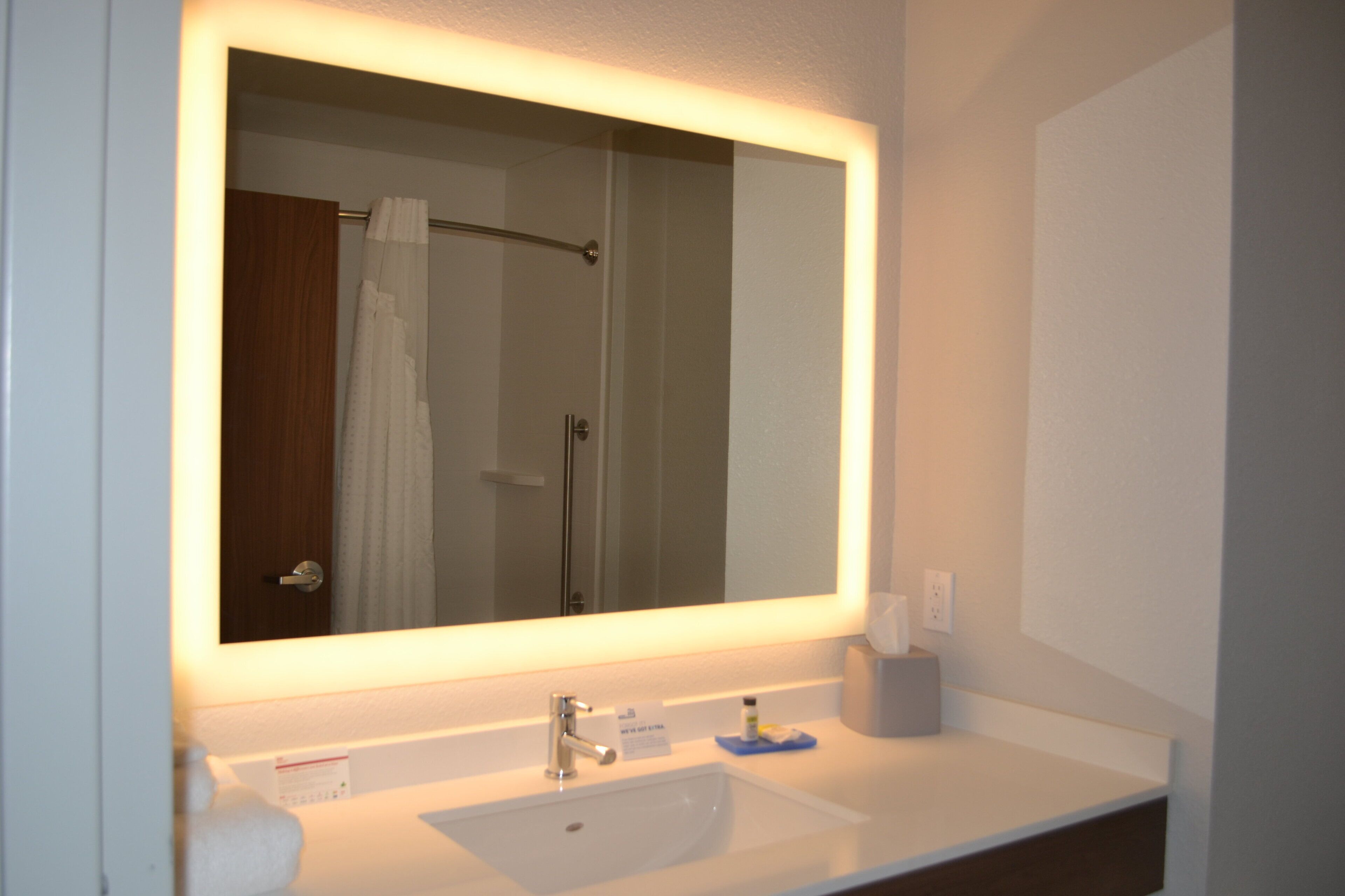 A bathroom with a large mirror, a white sink, a towel rack, and a shower visible in the reflection.