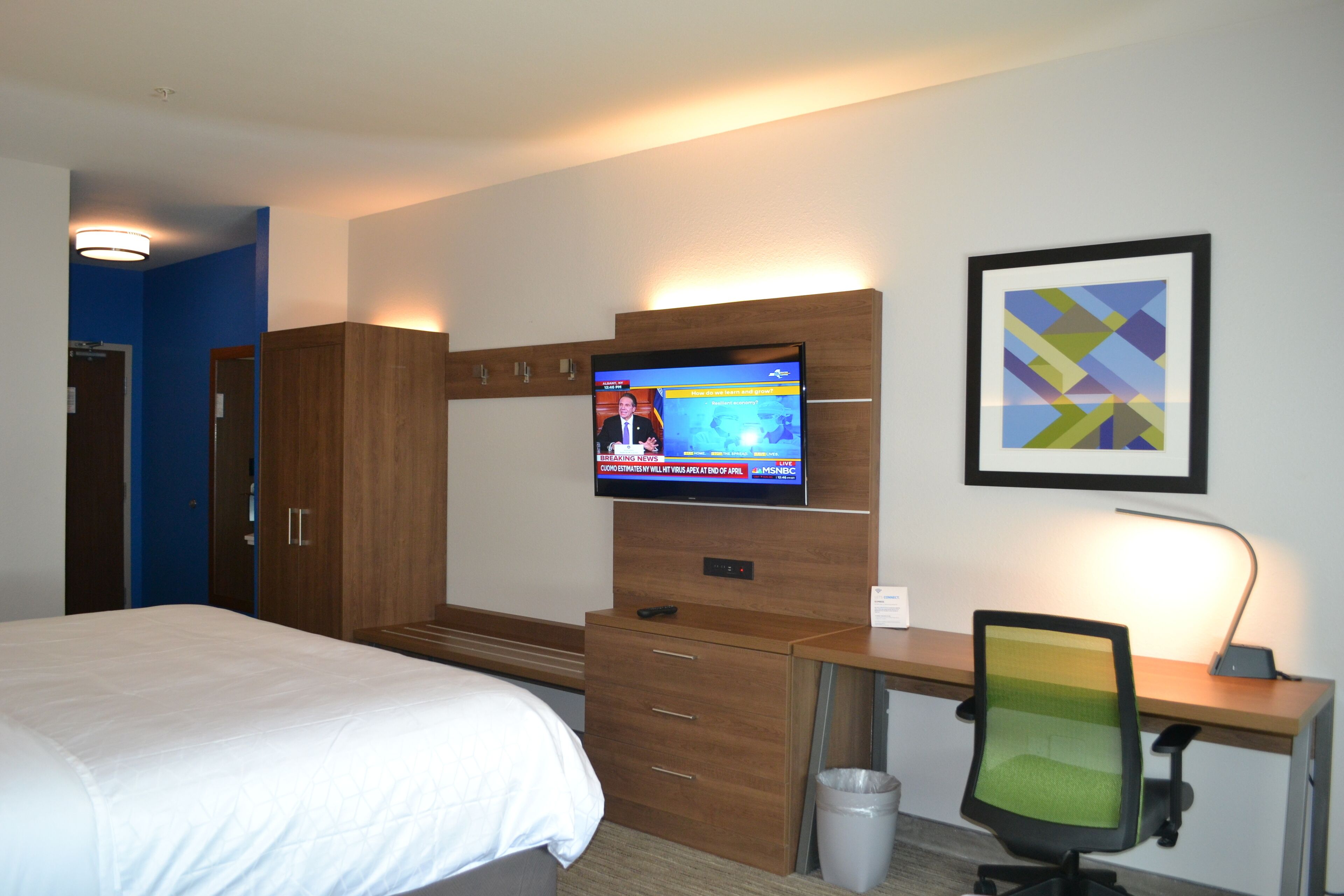 A hotel room with a bed, a desk with a computer, a TV on the wall, and a trash can.