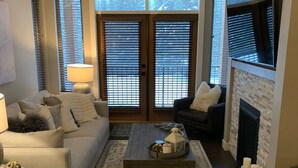 Ein Wohnzimmer mit einer Couch, einem Couchtisch und einem Kamin. Zwei Fenster mit Jalousien, eine Lampe und ein Teppich sind ebenfalls vorhanden.